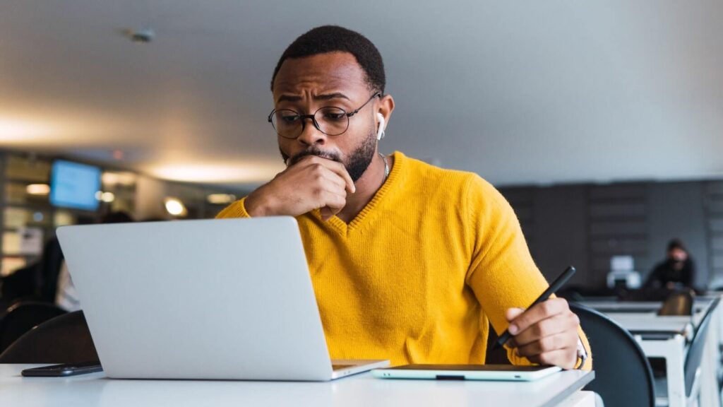 A man in glasses wearing a yellow sweater, looking at a laptop screen and thinking.