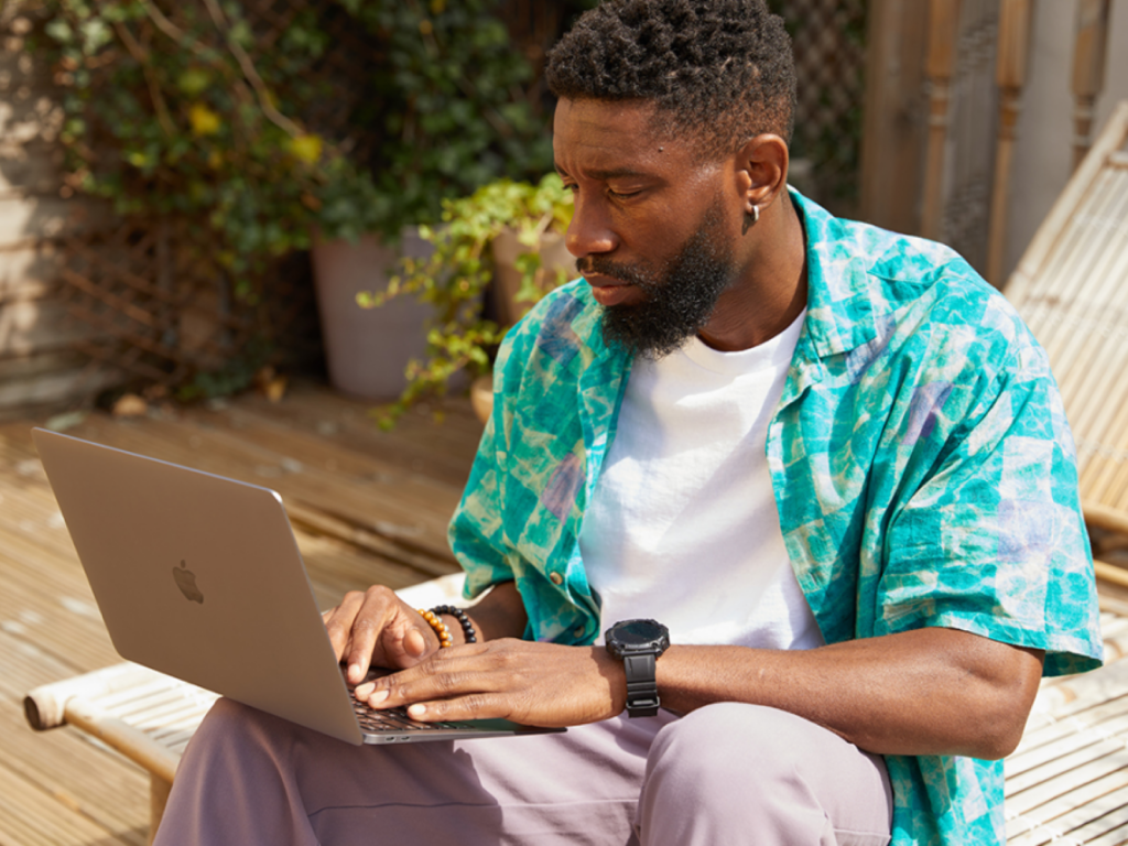Man in a colorful shirt focuses on a laptop while sitting on a sunlit deck. He appears thoughtful.