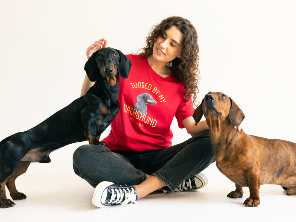 A woman in a red shirt with a dachshund graphic sits cross-legged, smiling at two dachshunds.