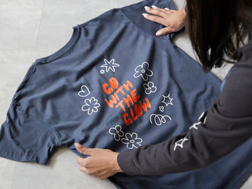 A person arranges a navy t-shirt with the phrase "Go With the Glow" in orange, surrounded by white doodle flowers and stars.