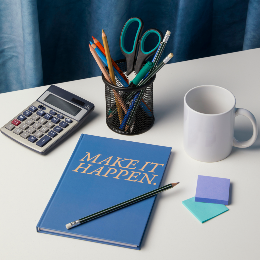 A custom "Make It Happen" notebook, pencil, calculator, coffee mug, notepad, pen, and notebook arranged on a white table.