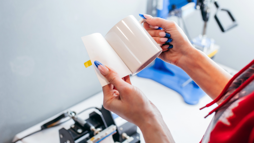 A person with blue-painted nails holds a plain white mug and a piece of paper, likely for customization, in a workshop with equipment in the background.