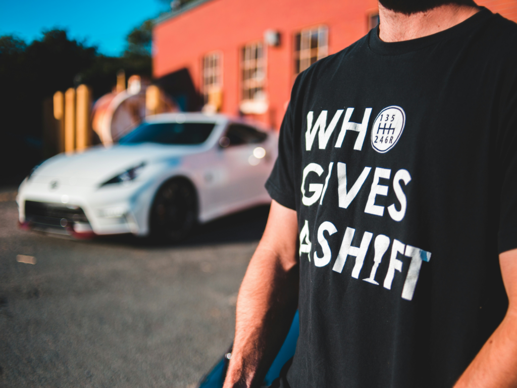 A man wearing a black “Who gives a shift” t-shirt with a blurred supercar in the background.