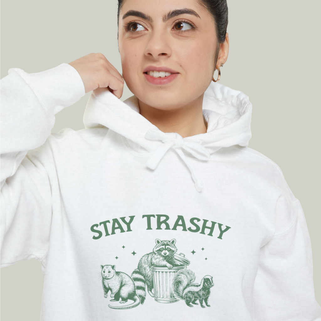 A close-up shot of a woman wearing a white “Stay Trashy” hoodie with an illustrated raccoon and other animals.