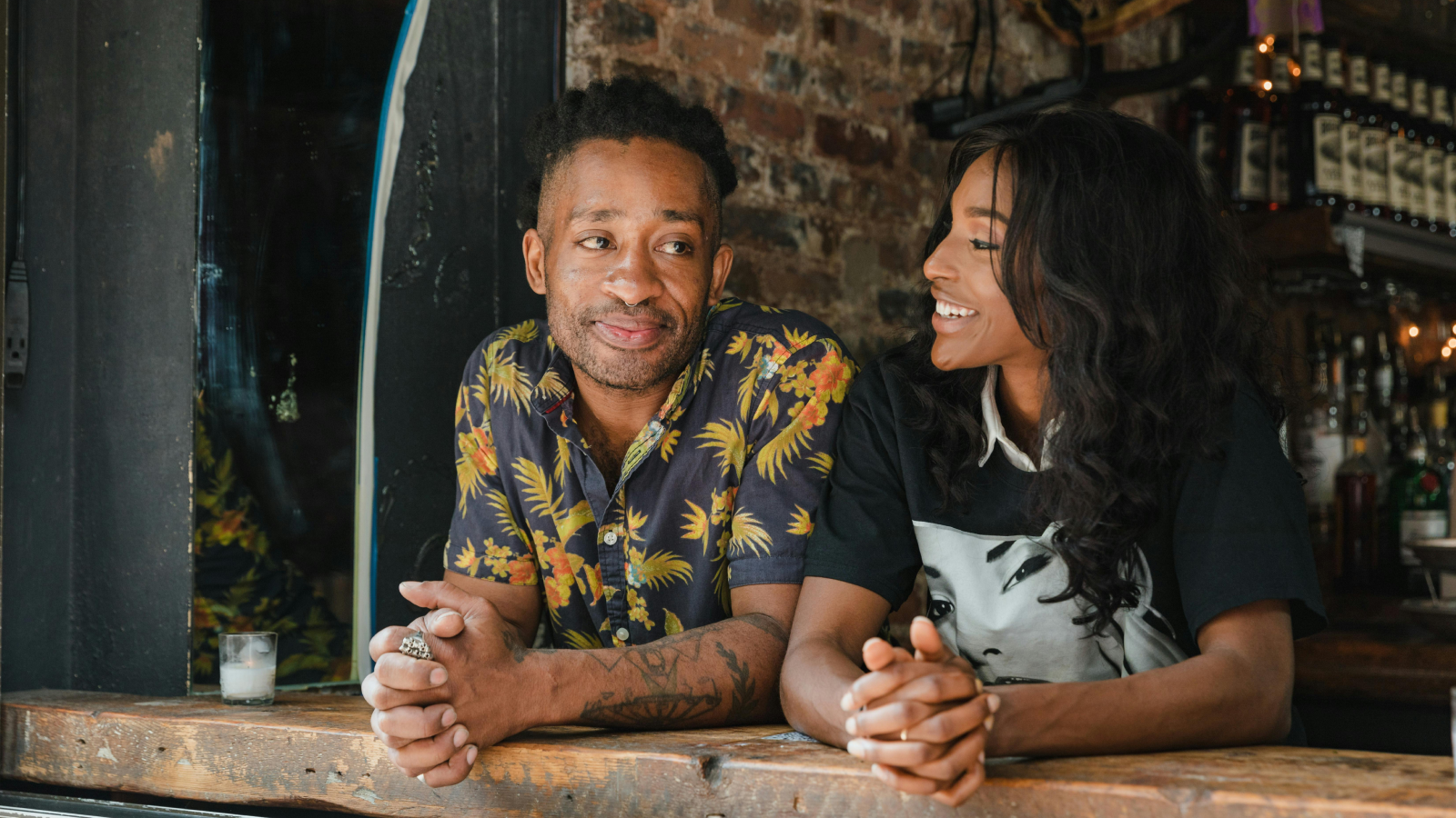 A man in a vibrant all-over-print shirt and a woman sit at a bar, engaged in friendly conversation.
